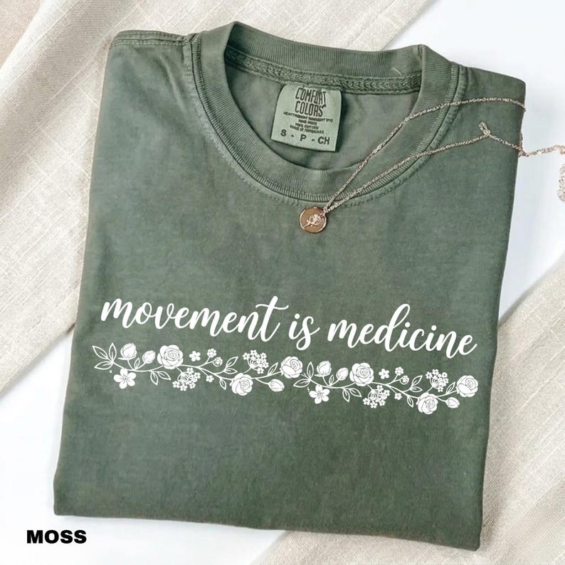 K&ouml;nnte beinhalten: Ein moosgr&uuml;nes T-Shirt mit dem wei&szlig;en Schriftzug "movement is medicine" und einem floralen Design. Eine silberne Halskette mit Anh&auml;nger liegt auf dem T-Shirt, das auf einer neutralen Oberfl&auml;che gefaltet ist.