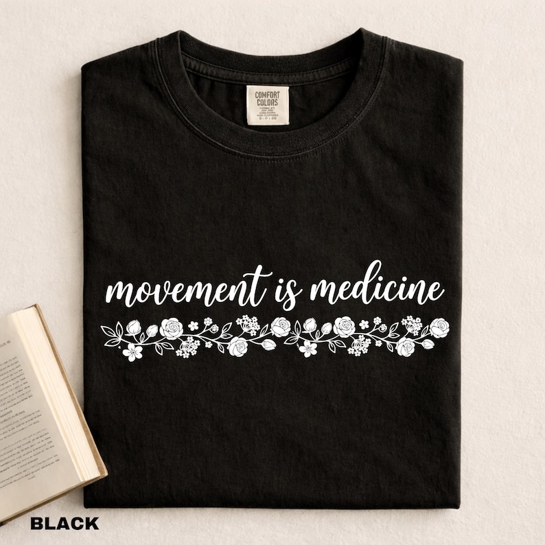 K&ouml;nnte beinhalten: Schwarzes T-Shirt mit dem wei&szlig;en Schriftzug "movement is medicine" und einem floralen Design. Das T-Shirt ist gefaltet. Das Wort "BLACK" steht unten. Ein Buch ist links sichtbar.