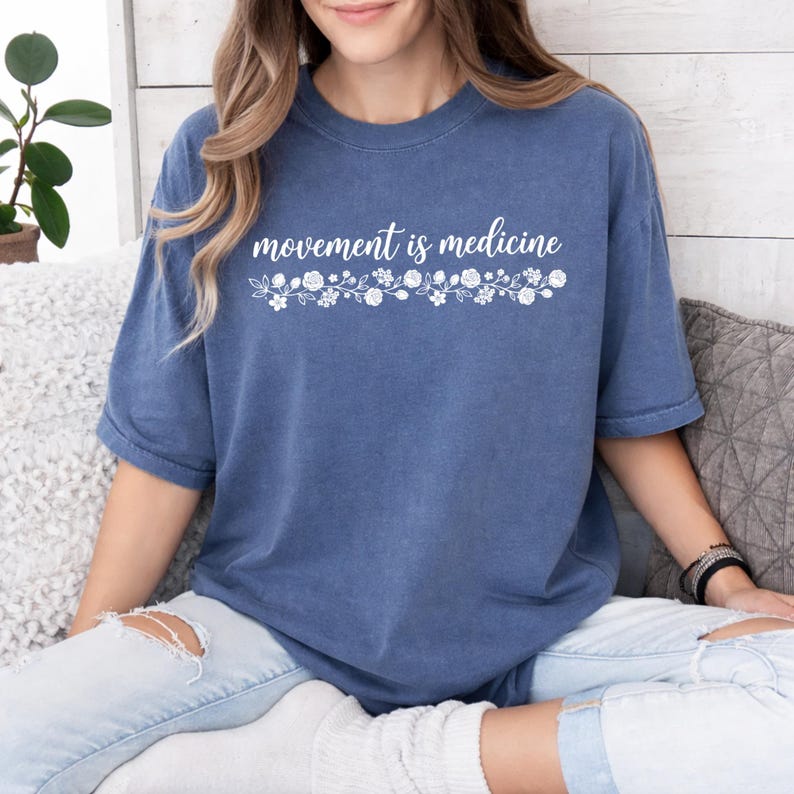 K&ouml;nnte beinhalten: Blaues T-Shirt mit dem wei&szlig;en Schriftzug "movement is medicine" und einem floralen Muster. Das kurz&auml;rmelige Shirt wird mit hellblauen Jeans getragen. Das Model sitzt auf einem Sofa.