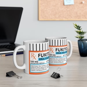 May include: Two white ceramic mugs with a prescription label design. The label reads "FUKIT Oral Solution 100 MG Unlimited Refills" with directions for stress, anxiety, and family time. The mugs are on a light wood surface.