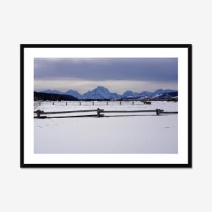May include: A framed landscape photograph of a snowy field with a wooden fence in the foreground. Mountains are visible in the distance under a cloudy sky. The photograph is framed in a black border.