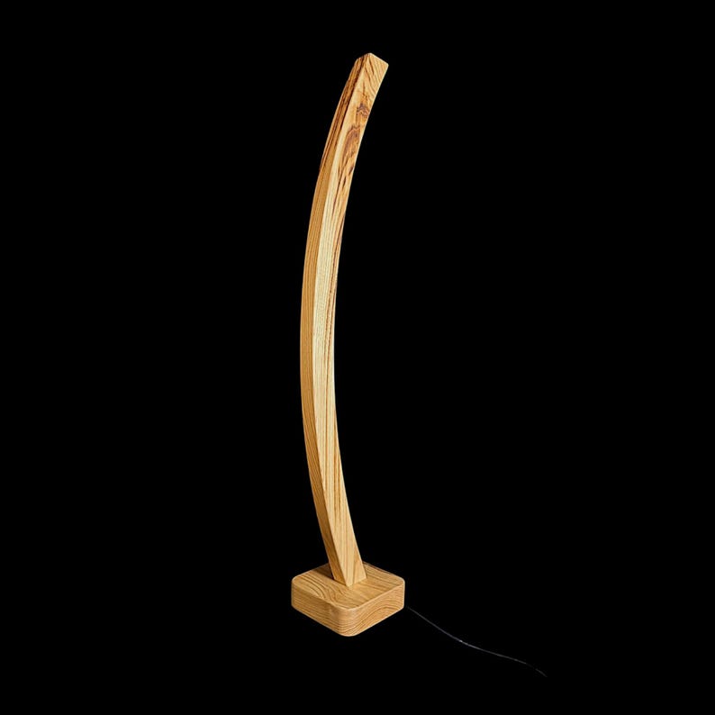 Op de afbeelding: Een moderne houten vloerlamp met een gebogen, slank ontwerp. De lamp heeft een lichtgekleurd, gebogen houten frame dat aan een vierkante basis is bevestigd. De lamp is gemaakt van hout en heeft een zwart netsnoer.