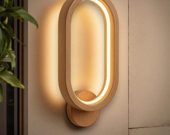 Puede incluir: Aplique de pared ovalado de madera clara con una fuente de luz lineal. La lámpara presenta una forma ovalada con una base circular. El diseño es minimalista y elegante, ideal para interiores modernos. La iluminación es cálida.
