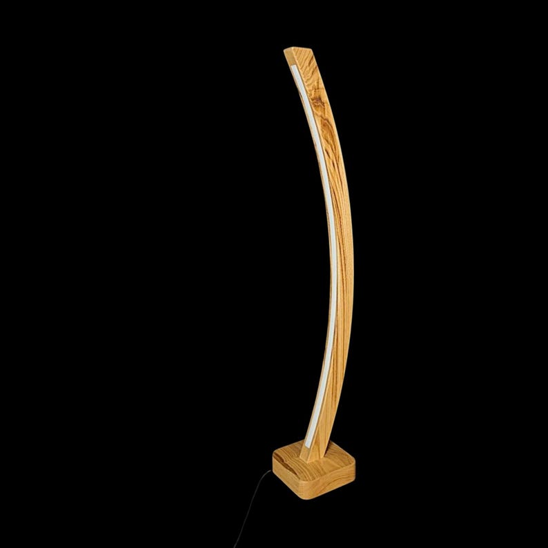 Op de afbeelding: Een moderne, gebogen vloerlamp van lichtkleurig hout met een natuurlijke nerf. De lamp heeft een witte LED-lichtstrip langs de binnenbocht. De basis is een vierkant blok van bijpassend hout. Een zwart netsnoer komt uit de basis. Hoogte: circa 150 cm.