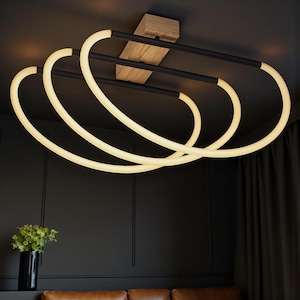 Puede incluir: Luminaria de techo moderna con base de madera y tres anillos de luz ovalados interconectados y brillantes. Los anillos de luz son blancos y la luminaria tiene detalles en negro. La luminaria está montada en un techo oscuro.