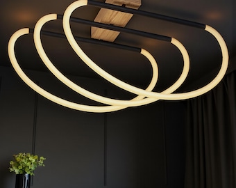 May include: Modern ceiling light fixture with a wooden base and three interconnected, glowing, oval-shaped light rings. The light rings are white and the fixture has black accents. The fixture is mounted on a dark ceiling.
