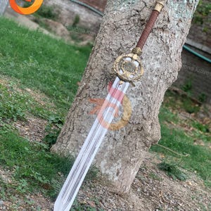 Puede incluir: Una espada decorativa con una hoja plateada y una empuñadura dorada ornamentada. El mango está envuelto en cuero marrón. La espada está apoyada contra un tronco de árbol. La hoja mide unos 76 cm de largo.