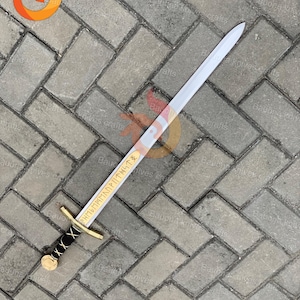 May include: A silver-bladed sword with a gold crossguard and a black-wrapped handle. The blade has gold lettering. The sword is lying on a gray brick surface. The sword is approximately 30 inches long.