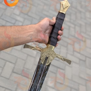 May include: A medieval-style sword with a black blade and gold-colored hilt and crossguard. The handle is wrapped in black material. The sword is held in a hand. The brand name "Bauona knives crafts" is visible in the background.