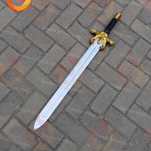 May include: A decorative sword with a silver blade and a gold-coloured hilt. The handle is black with gold accents. The sword is lying on a grey brick surface.