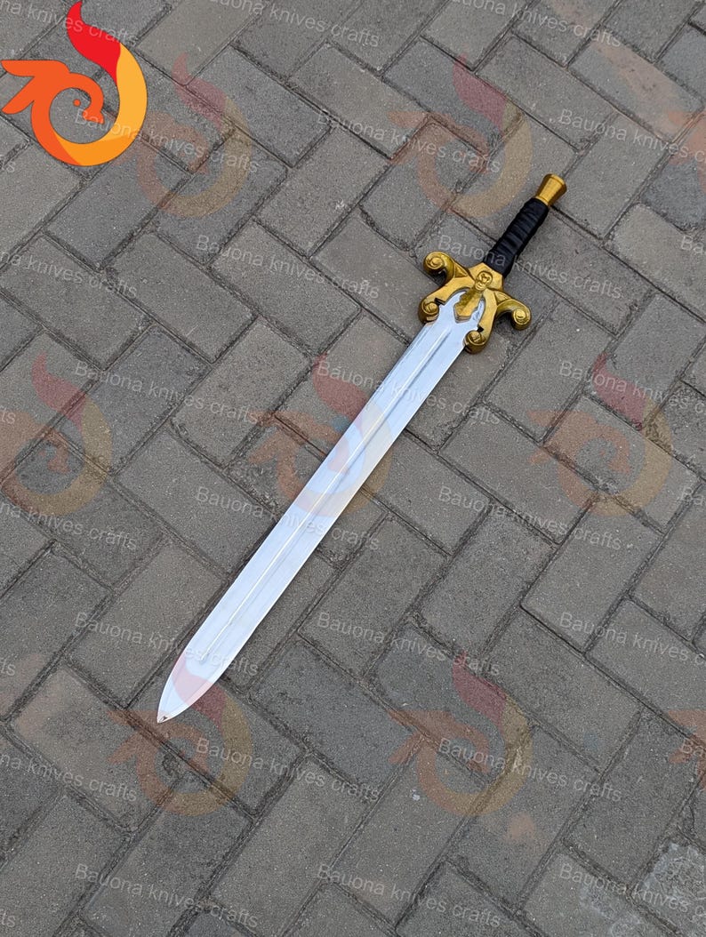 May include: A decorative sword with a silver blade and a gold-coloured hilt. The handle is black with gold accents. The sword is lying on a grey brick surface.