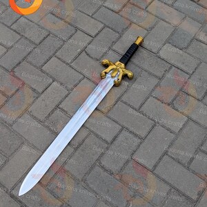 May include: A decorative sword with a silver blade and a gold-coloured hilt. The handle is black with gold accents. The sword is lying on a brick surface.
