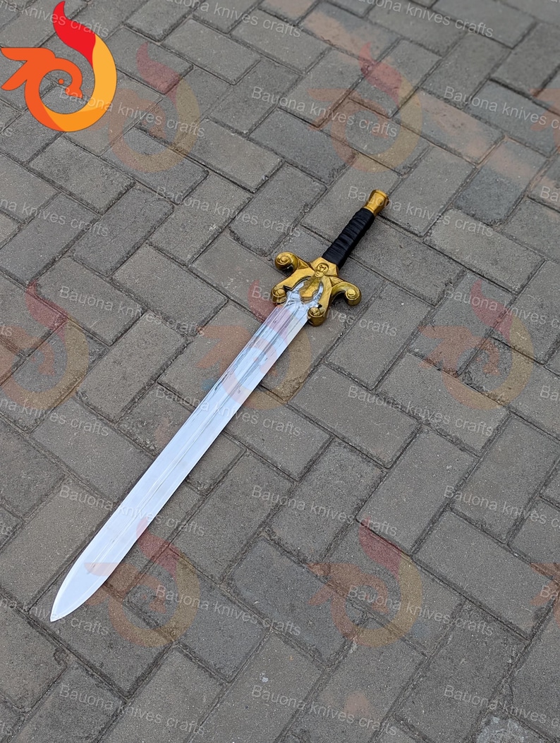 May include: A decorative sword with a silver blade and a gold-coloured hilt. The handle is black with gold accents. The sword is lying on a brick surface.