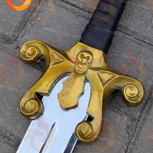 May include: A detailed view of a decorative sword. The hilt is gold-coloured with a skull motif, and the blade is silver. The handle is wrapped in black. This item is likely for display or collection.