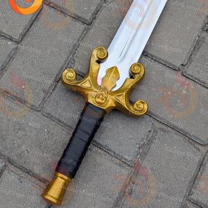 May include: A decorative sword with a gold-coloured hilt and a silver blade. The hilt features intricate designs and a black wrapped handle. The sword is displayed on a textured surface.