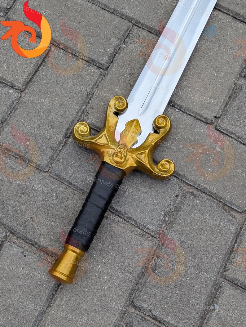 May include: A decorative sword with a gold-coloured hilt and a silver blade. The hilt features intricate designs and a black wrapped handle. The sword is displayed on a textured surface.