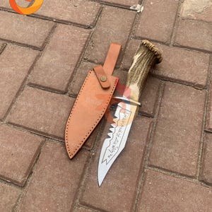 May include: A hunting knife with a polished steel blade and a handle made of antler. The knife is accompanied by a brown leather sheath. The blade has an etched design and text. The knife is laying on a brick surface.