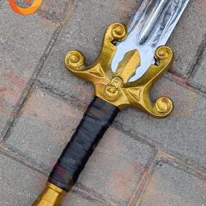 May include: A decorative sword with a polished silver blade and a gold-coloured hilt. The hilt features an ornate design with a black wrapped handle. The sword is likely a display piece or costume accessory.