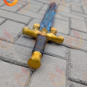 May include: A decorative sword with a gold-coloured hilt and pommel. The blade has a blue and silver finish. The handle is wrapped in dark blue and brown material. The sword is lying on a grey stone surface.