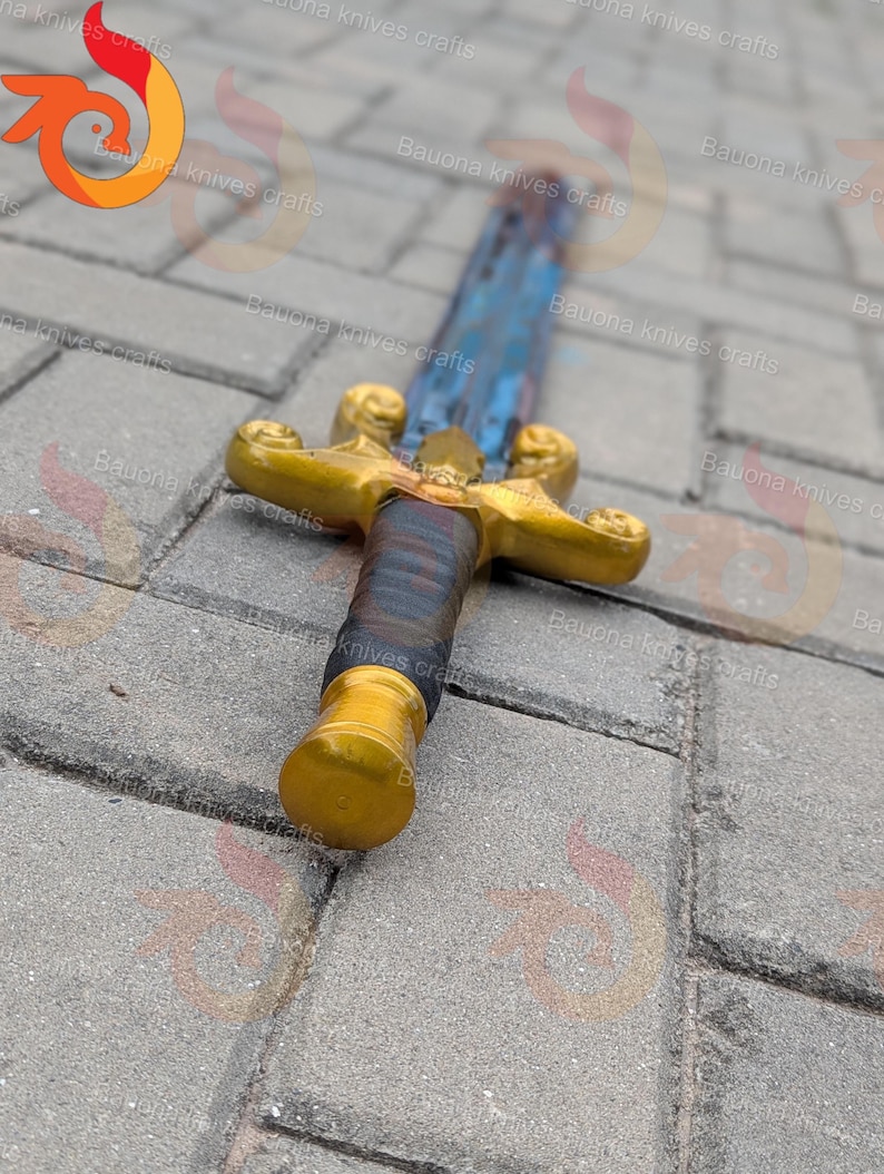 May include: A decorative sword with a gold-coloured hilt and pommel. The blade has a blue and silver finish. The handle is wrapped in dark blue and brown material. The sword is lying on a grey stone surface.