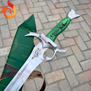 May include: A silver metal sword with a green handle featuring black decorative patterns and a silver pommel. The blade displays a small etched feather symbol and the text "lighter than a feather". A green leather sheath with brown straps is included.