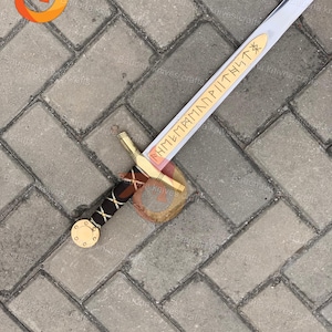 May include: A silver and gold sword with a brown and black handle. The blade has a gold inscription. The sword is lying on a gray brick surface. The sword is a replica of a historical weapon.