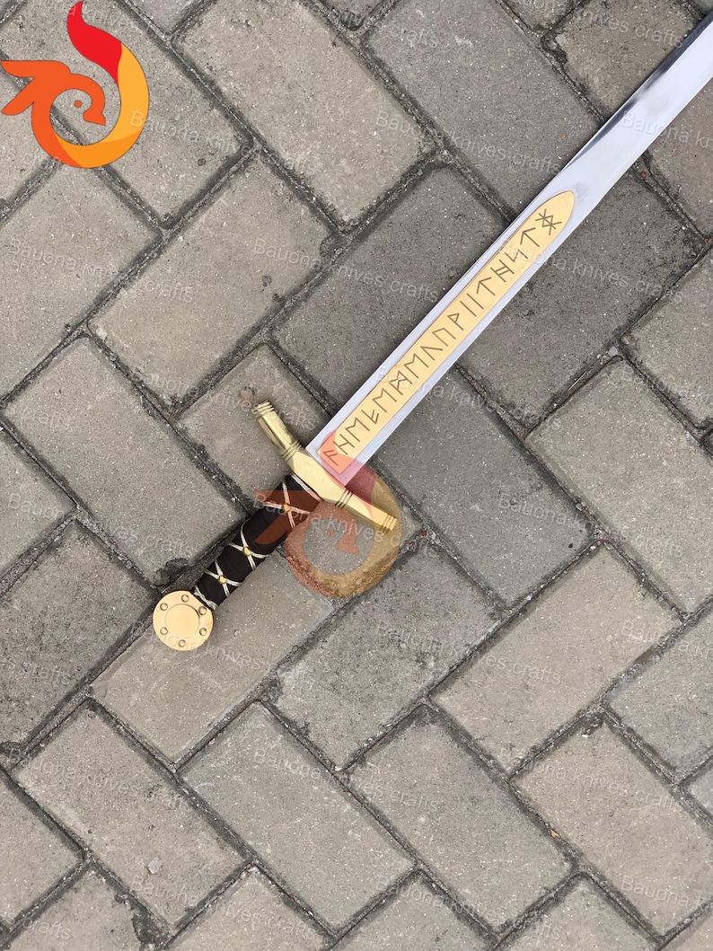 May include: A silver and gold sword with a brown and black handle. The blade has a gold inscription. The sword is lying on a gray brick surface. The sword is a replica of a historical weapon.