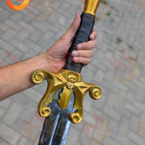 May include: A gold and silver sword with a skull design on the crossguard. The handle is wrapped in black material. The blade is silver and the hilt is gold. The sword is held in a hand.