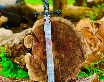 Puede incluir: Una espada decorativa con una hoja estampada y un mango negro, apoyada sobre un tronco de madera. La espada tiene una guarda y una empuñadura detallada. La hoja tiene un diseño único. El fondo es un entorno natural con follaje verde.