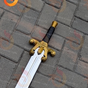May include: A decorative sword with a gold-coloured hilt featuring a skull design and a black leather-wrapped handle. The blade is silver and the sword is displayed on a brick surface.