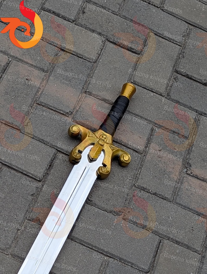 May include: A decorative sword with a gold-coloured hilt featuring a skull design and a black leather-wrapped handle. The blade is silver and the sword is displayed on a brick surface.