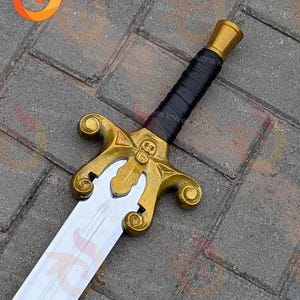 May include: A decorative sword with a gold hilt and a silver blade. The hilt has a skull design and a black leather-wrapped grip. The sword is displayed on a grey brick surface.