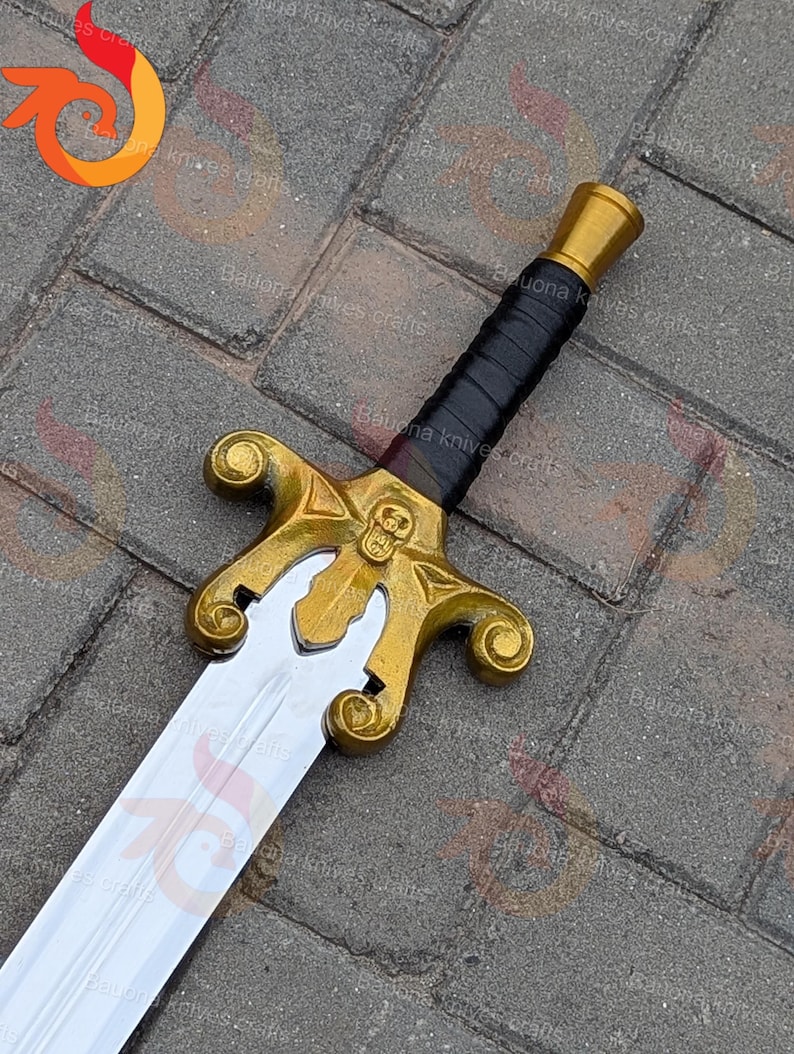 May include: A decorative sword with a gold hilt and a silver blade. The hilt has a skull design and a black leather-wrapped grip. The sword is displayed on a grey brick surface.