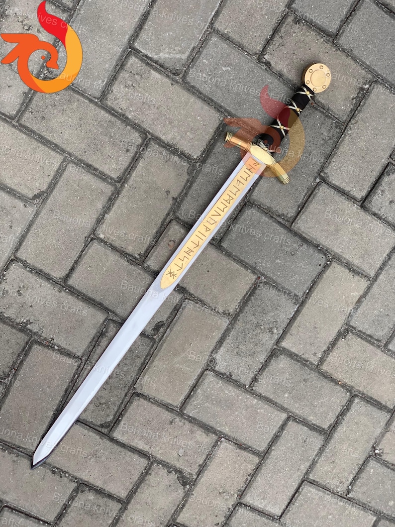 May include: A decorative sword with a silver blade and gold accents. The hilt features a black grip with gold detailing and a circular gold pommel. The blade has gold lettering. The sword is displayed on a gray brick surface.