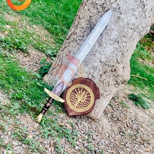 May include: A decorative sword and shield set. The sword has a silver blade, a brown and gold handle, and a gold crossguard. The shield is brown with a gold emblem. The set is displayed outdoors near a tree.