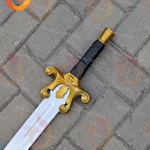 May include: A decorative sword with a gold-coloured hilt and a silver blade. The hilt features a skull design and ornate detailing. The handle is wrapped in black material. The sword is displayed on a grey brick surface.