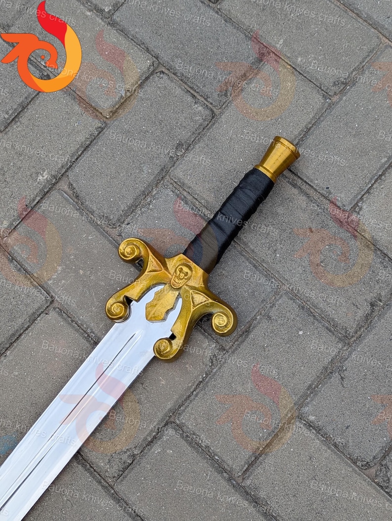 May include: A decorative sword with a gold-coloured hilt and a silver blade. The hilt features a skull design and ornate detailing. The handle is wrapped in black material. The sword is displayed on a grey brick surface.