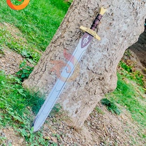 Può includere: Una spada argentata con un manico in pelle marrone e un pomo dorato. La spada è appoggiata contro un tronco d'albero. La spada ha una guardia a forma di croce e l'iscrizione "The Crusader" sulla lama.