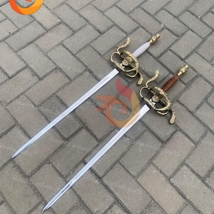 May include: Two decorative swords with ornate gold-colored hilts and long, silver blades. One sword has a white handle, the other a brown handle. The swords are displayed on a gray brick surface.