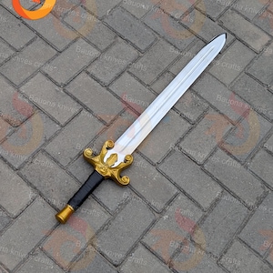 May include: A decorative sword with a silver blade and a gold-coloured hilt. The hilt features an ornate design with a black grip. The sword is displayed on a grey brick surface.