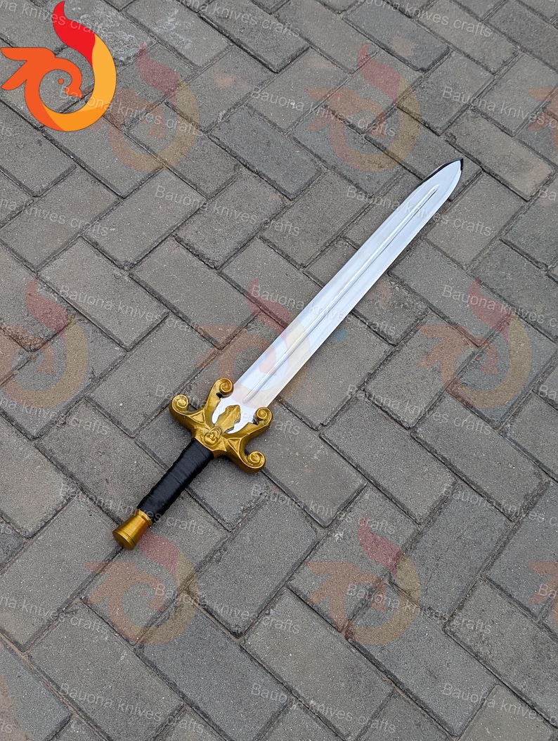 May include: A decorative sword with a silver blade and a gold-coloured hilt. The hilt features an ornate design with a black grip. The sword is displayed on a grey brick surface.