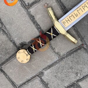 May include: A decorative sword with a gold handle and a black grip adorned with gold accents. The blade is silver with the word "FHMEMAR" engraved in gold. The sword's pommel is a gold disc.