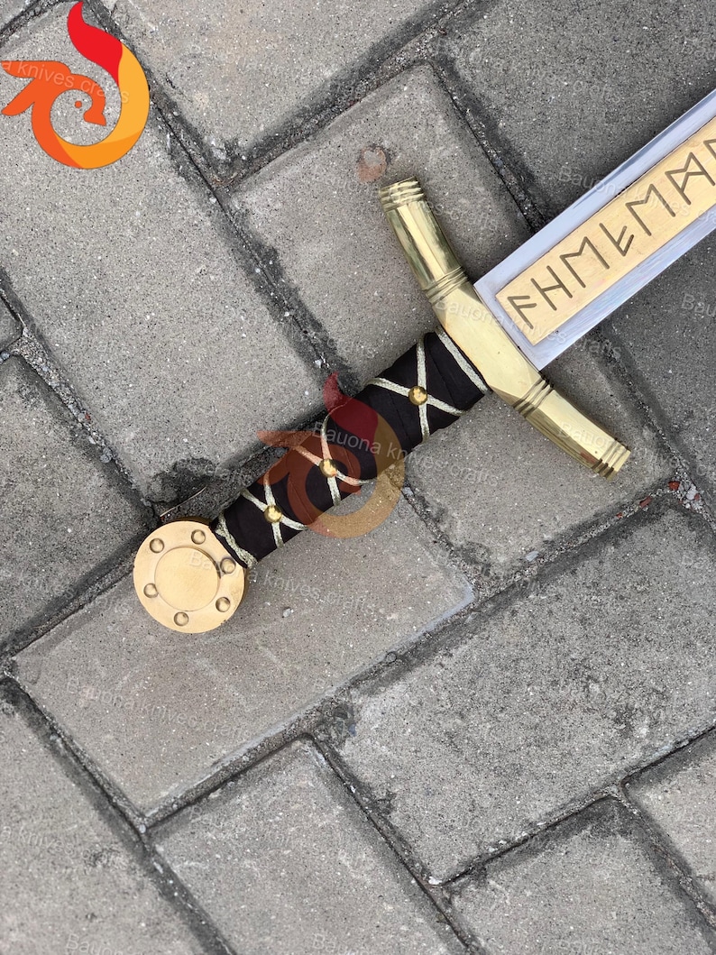May include: A decorative sword with a gold handle and a black grip adorned with gold accents. The blade is silver with the word "FHMEMAR" engraved in gold. The sword's pommel is a gold disc.