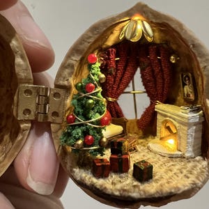 May include: A detailed miniature Christmas scene housed within a walnut shell. Features include a small Christmas tree with red and gold baubles, a fireplace, and tiny presents. Red curtains frame a window. The scene is illuminated with small lights.