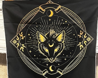 May include: Black tapestry featuring a golden fox design within a geometric frame. The fox has pointed ears and a crescent moon on its forehead. The design includes stars, moons, and floral elements. The tapestry has a white border.