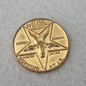 May include: A gold-coloured circular coin with a pentagram design. The centre features a goat head within the star. The coin is inscribed with the words "IN GOD WE'RE DAMNED", "SERPENT", "DRAGON", "LUCIFER", "MORNING STAR", and "SATAN".