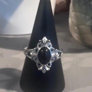 Peut inclure: Bague en argent ornée d'une pierre noire ovale. La bague présente un motif filigrané orné autour de la pierre. La bague est présentée sur un présentoir noir en forme de cône.