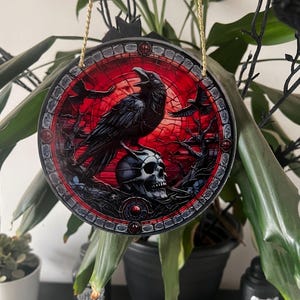 May include: Round, gothic-style ornament featuring a black raven perched on a skull against a red stained-glass background. The ornament is trimmed with a decorative border and suspended by a gold-colored cord. The design includes additional ravens and dark, gothic elements.