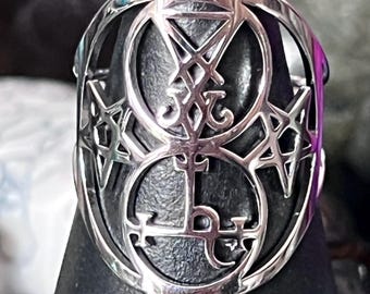 May include: A silver-colored ring featuring an intricate design of overlapping circles, stars, and symbols. The ring is displayed on a black stand, showcasing its detailed craftsmanship and gothic aesthetic. The ring's design is symmetrical and detailed.