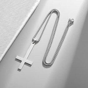 Op de afbeelding: Zilverkleurige ketting met een omgekeerde kruis hanger. De hanger is een eenvoudig, plat ontwerp met een lange verticale balk en een horizontale balk. De ketting is een box chain stijl. De ketting wordt getoond op een lichtgrijze ondergrond.